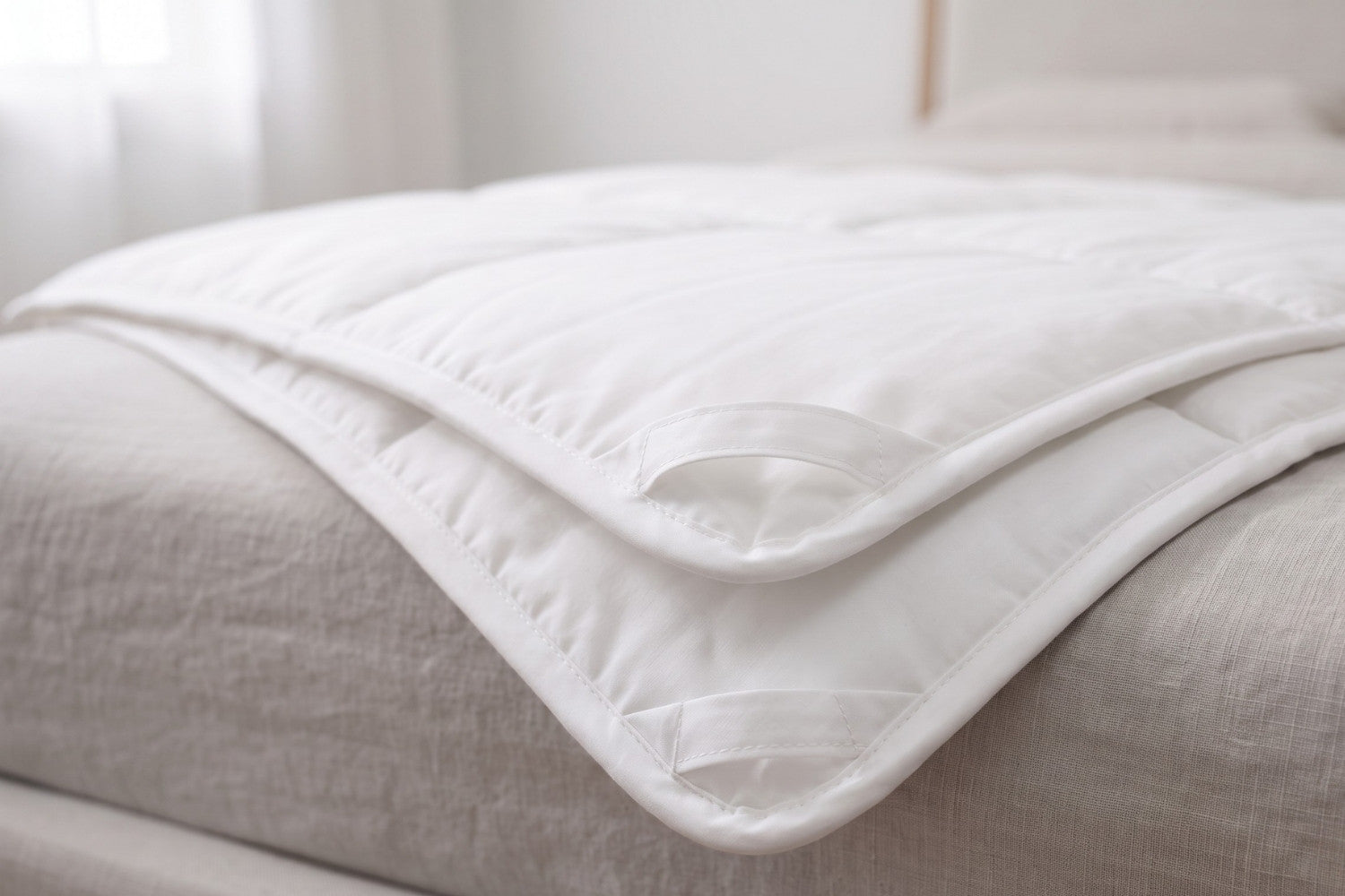A close-up of a white Wisewool Duvet showing off its corner loops, draped over a bed with light beige bedding in a softly lit bedroom.