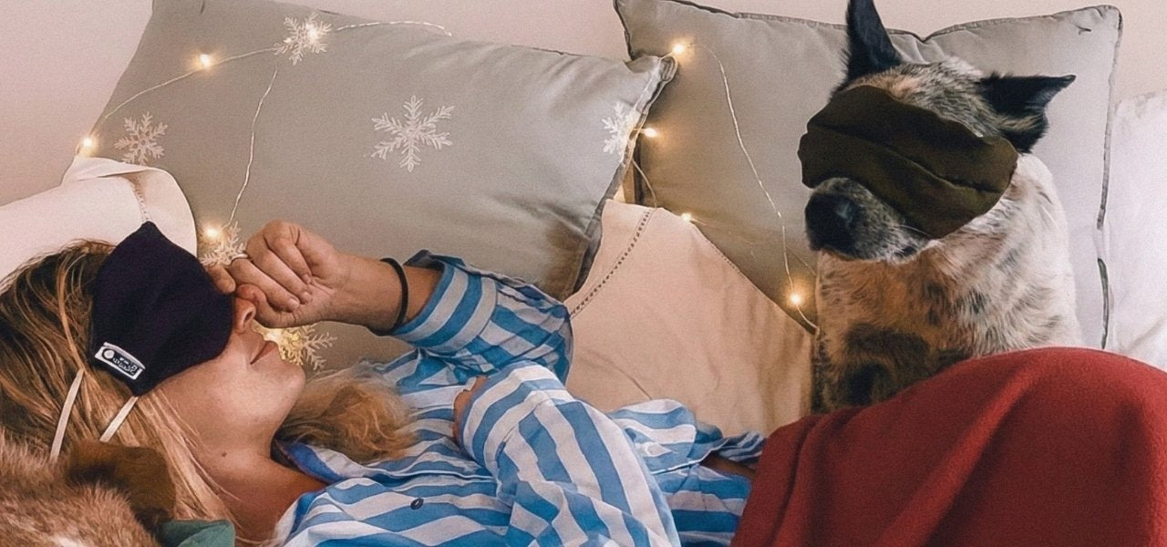 Sam and her dog lying on a couch with eye masks, surrounded by soft lighting and pillows.